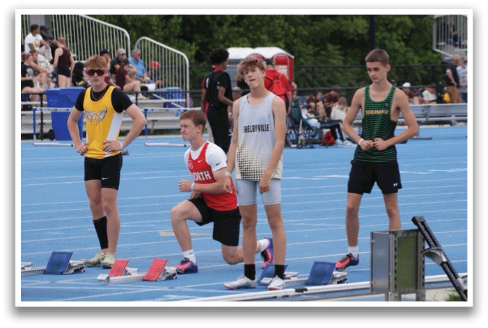 A group of young men are standing on a track, wearing shorts and shirts. They are lined up and appear to be waiting for a race to start. Contenido generado con IA