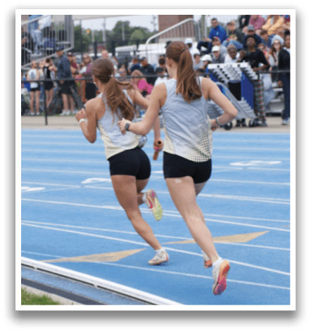 Two female runners are racing on a track, with one ahead of the other. They are wearing shorts and running shoes. A crowd of people is watching the race from the stands. Contenido generado con IA
