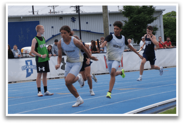 Three young boys are running on a track, with one of them wearing a green shirt. They are all running towards the finish line, with one of them leading the pack. There are several other people in the background, watching the race. Contenido generado con IA