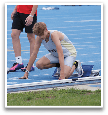 A man is running on a track, wearing a blue shirt and white shorts. He is running in front of a crowd of people, some of whom are sitting on chairs. The spectators are watching the man as he runs, possibly in a competition. Contenido generado con IA