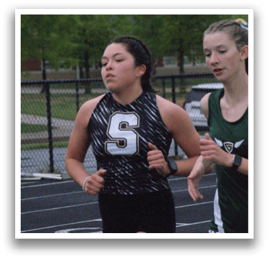 Two female runners are on a track, one wearing a green and white outfit and the other wearing a black and white outfit. They are running in opposite directions. Contenido generado con IA