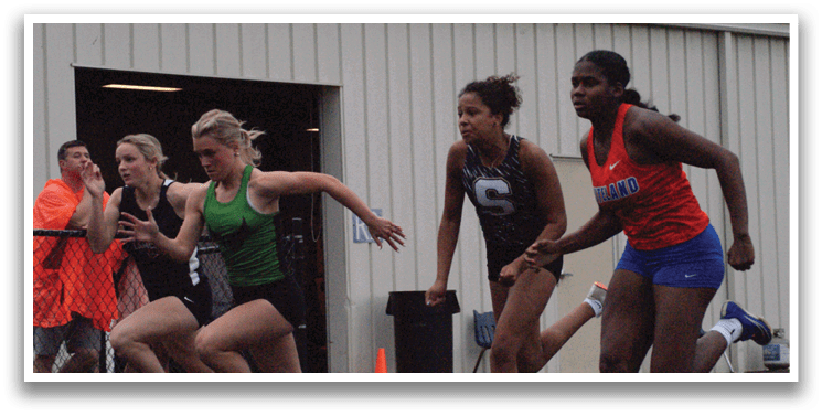 Three female runners are racing on a track, with one wearing a green shirt and the other two wearing red. Contenido generado con IA