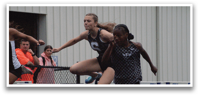 Two female athletes are running on a track, one of them jumping over a hurdle. Contenido generado con IA