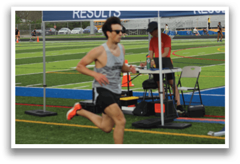 A man running on a track with a blue and white tent in the background. Description generated by AI