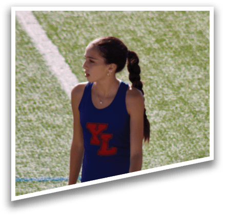 A girl wearing a blue and white shirt and shorts stands on a field. Description generated by AI