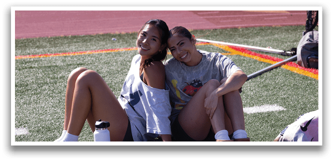 Two women are sitting on the ground, smiling and posing for a picture. They are surrounded by various items, including a backpack, a bottle, and a handbag. AI generated content
