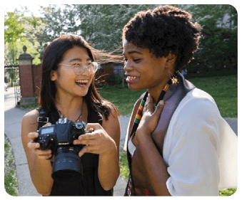 Two women are standing on a grassy field, one holding a camera and the other smiling. They are both wearing glasses. AI generated content
