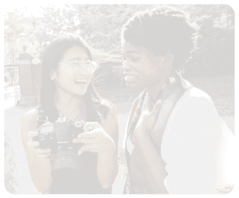 Two women are standing on a sidewalk, one holding a camera and the other smiling. They are both wearing glasses. AI generated content