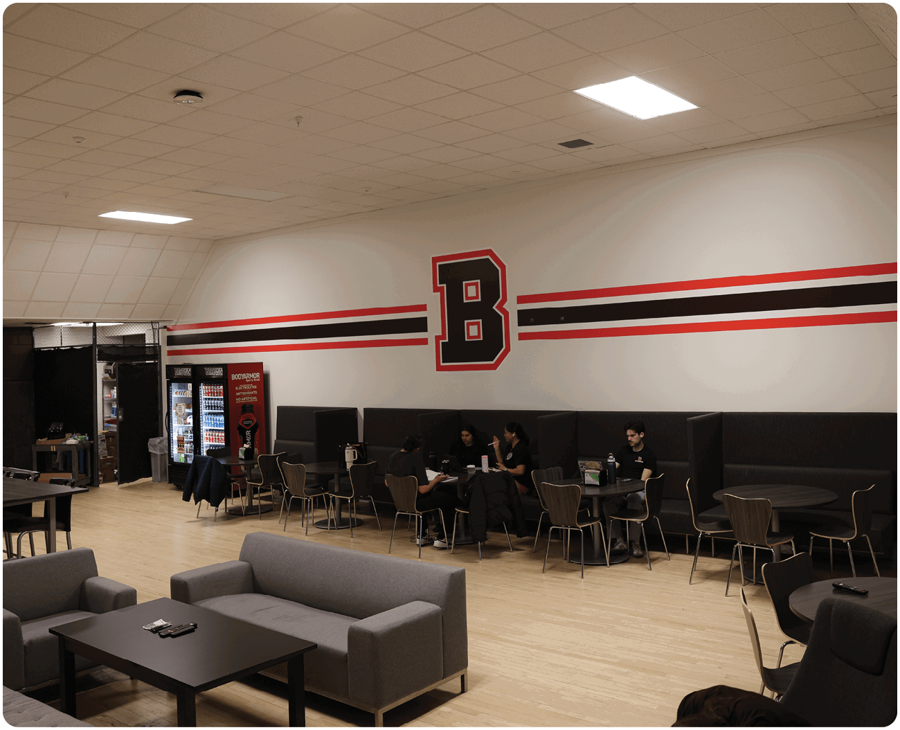 A group of people are sitting in a room with a large B on the wall. There are several couches and chairs in the room, and a dining table with a vase on it. A refrigerator is also present in the room. AI generated content