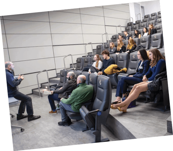 A group of people are sitting in a classroom, listening to a man speak. There are several chairs and a bench in the room. Some people are sitting on the bench, while others are sitting on chairs. The man is standing at the front of the room, giving a lecture. AI generated content