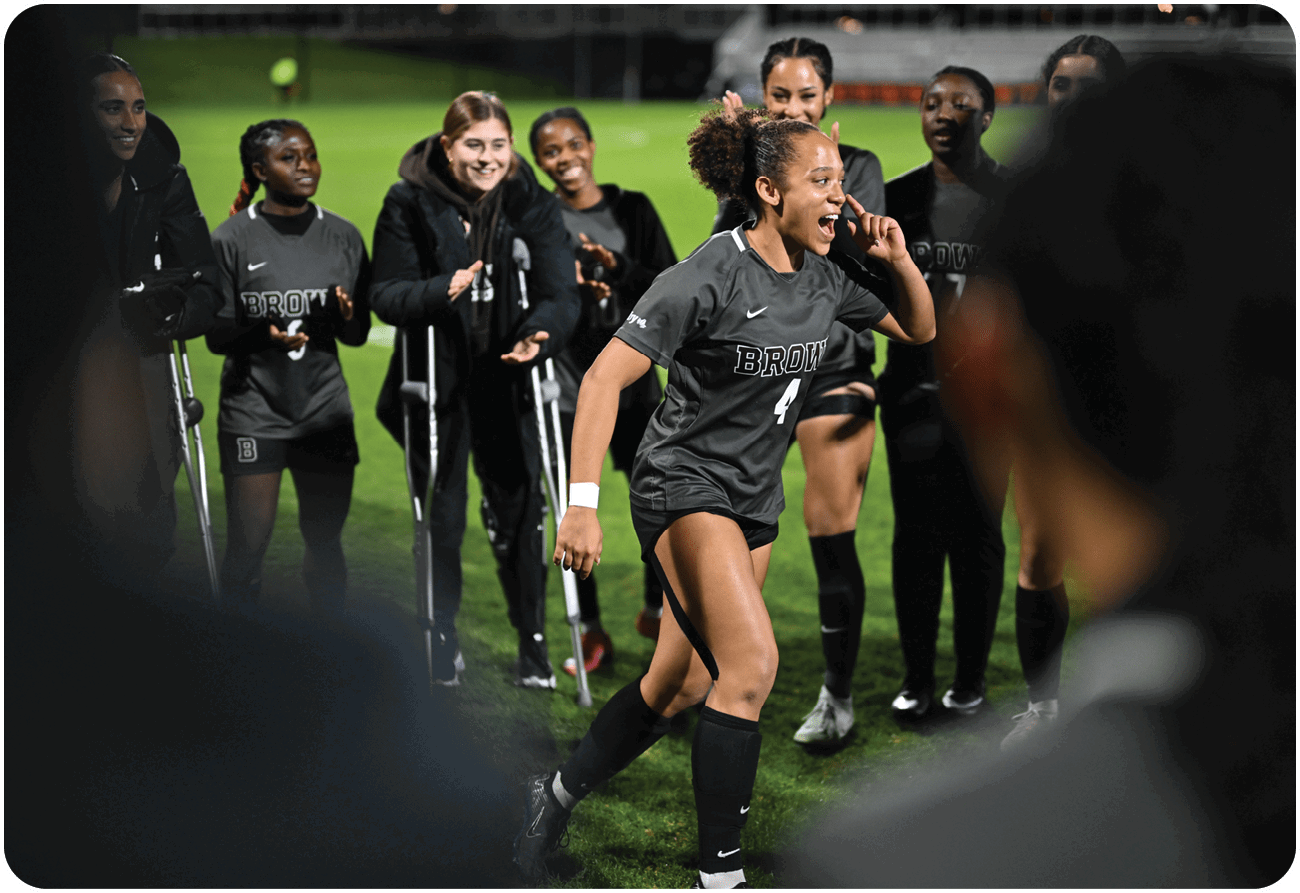 A group of people, including a woman in a grey shirt, are standing on a field. The woman is holding a cell phone to her ear, while the others are watching her. There are a few people wearing casts on their legs, and a tennis ball is visible in the scene. AI generated content