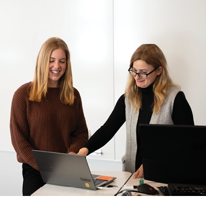 Two women are smiling and looking at a laptop. Description generated by AI