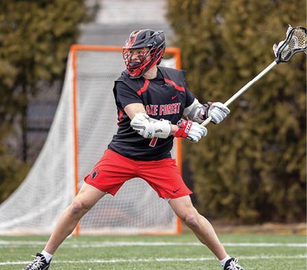 A man wearing a red and black shirt and red shorts is holding a lacrosse stick. Description generated by AI