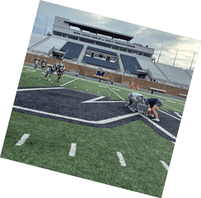 A sports stadium with empty blue seats and a group of people in sports uniforms standing on the field near a large letter M on a brick wall. Description generated by AI