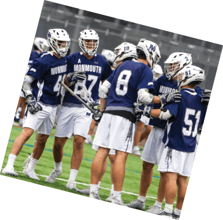 Four people wearing blue Monmouth lacrosse uniforms and white helmets are standing together on a field, holding lacrosse sticks. Description generated by AI