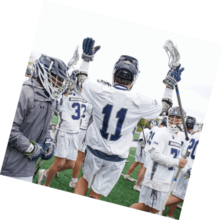 People wearing lacrosse gear are celebrating on a sports field with one person raising their arm and others gathered closely. Description generated by AI