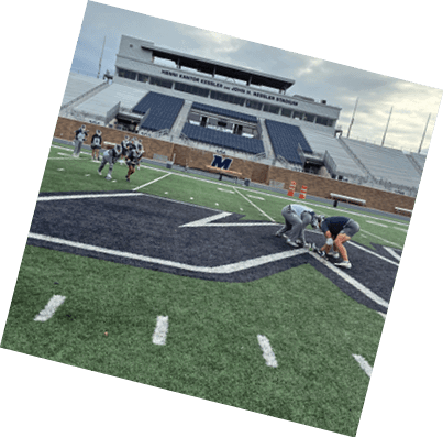 A sports stadium with empty blue seats and a group of people in sports uniforms standing on the field near a large letter M on a brick wall. Description generated by AI