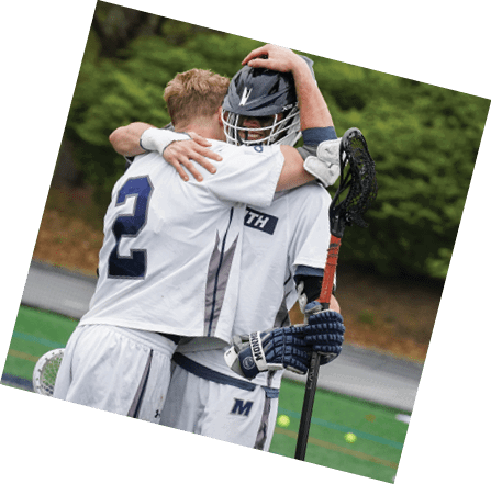 Two people in sports uniforms embrace on a field, one holding a lacrosse stick and wearing a helmet. Description generated by AI