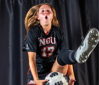A woman in a black and red soccer uniform is jumping in the air to kick a soccer ball. AI generated content