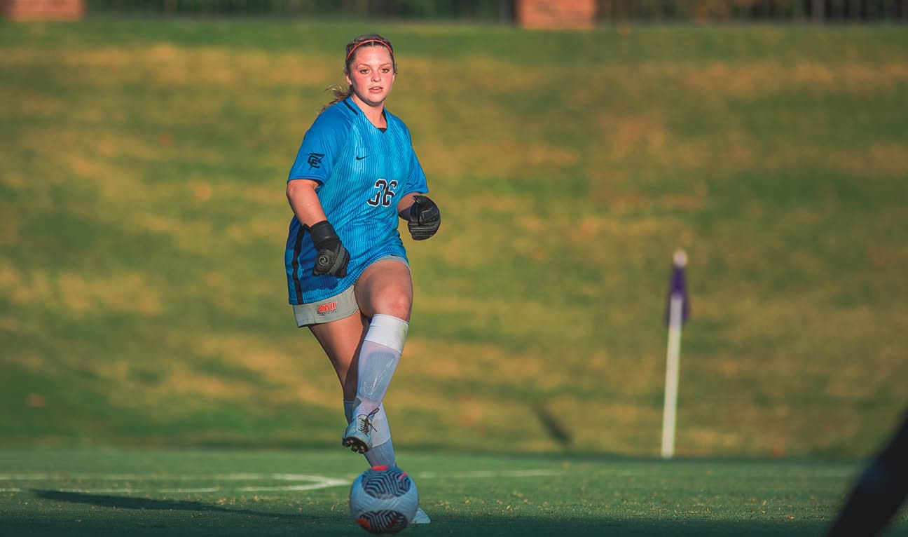 A woman in a blue shirt is running on a field, kicking a soccer ball. Another woman is running behind her. AI generated content