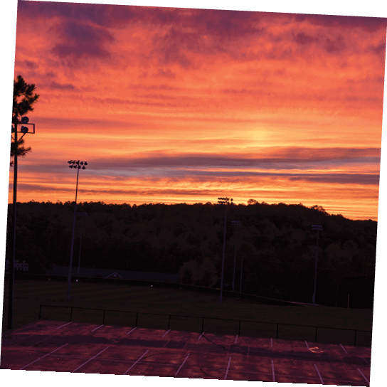 A sunset over a baseball field with a car parked in the parking lot. AI generated content