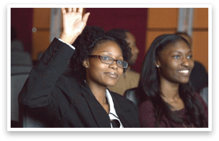 Two adults are seated indoors with one raising a hand and wearing glasses and a dark blazer while the other smiles and wears a dark top. Description generated by AI