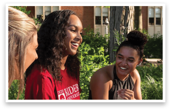 Three people are sitting outside on a sunny day, smiling and engaging in conversation, with one wearing a red Rider University shirt. Description generated by AI