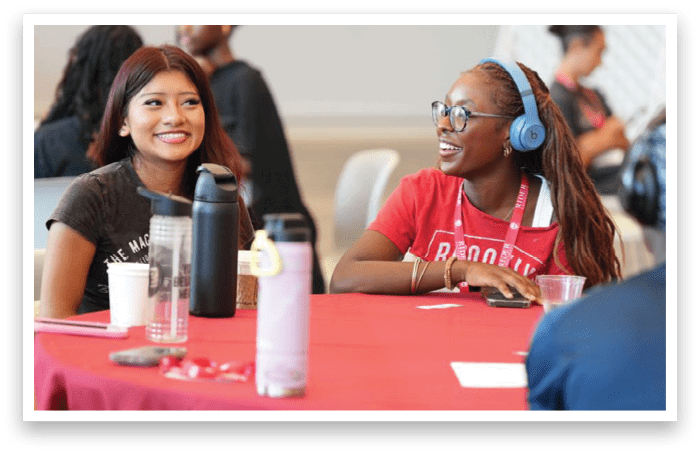 Two adults sit at a table covered with a red tablecloth, smiling and engaged in conversation, with water bottles and cups on the table. Description generated by AI