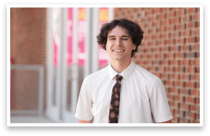 A person wearing a white short-sleeve shirt and a patterned tie is standing outdoors in front of a brick wall. Description generated by AI