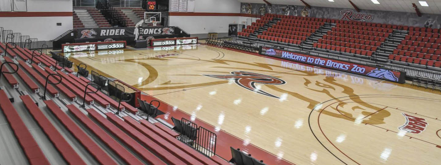 An empty indoor basketball court with red bleachers and a large Broncos logo at center court. Description generated by AI