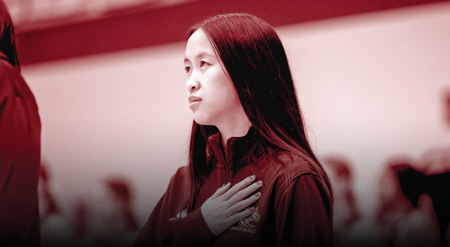 A person with long straight hair wearing a dark jacket stands with one hand over their chest in a solemn or respectful gesture. Description generated by AI