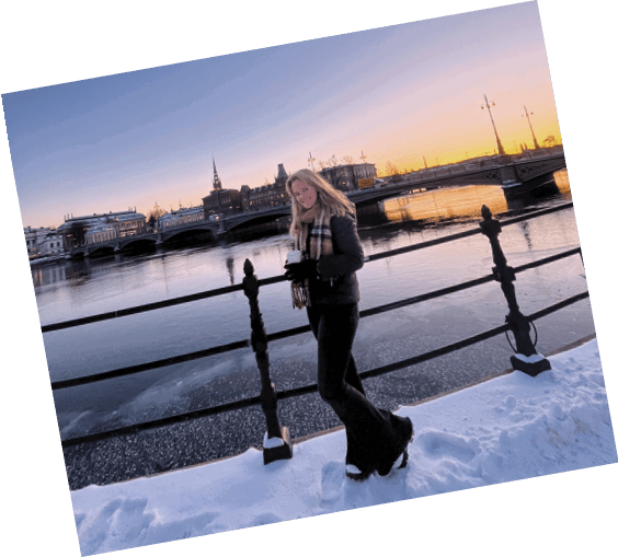 A woman in a black jacket and black pants stands on a bridge overlooking a body of water. She is holding a camera and smiling. AI generated content