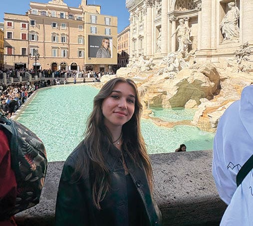 A woman is standing next to a fountain, wearing a green jacket. AI generated content