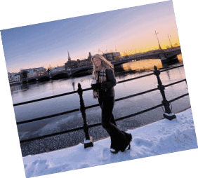 A woman in a black jacket and black pants stands on a bridge overlooking a body of water. She is holding a camera and smiling. AI generated content