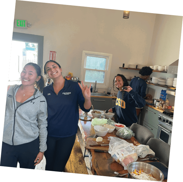 Two women are standing in a kitchen, smiling and waving at the camera. They are surrounded by various kitchen items such as a bowl, a spoon, a knife, and a cup. There is also a sink in the background. AI generated content