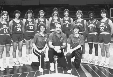 A group of adults wearing basketball uniforms and two adults in polo shirts pose together on an indoor basketball court. Description generated by AI