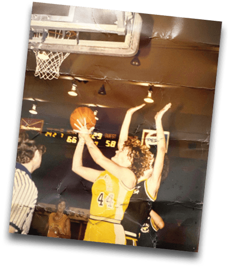Two people are reaching up near a basketball hoop as a basketball is about to go through the net in an indoor basketball court. Description generated by AI